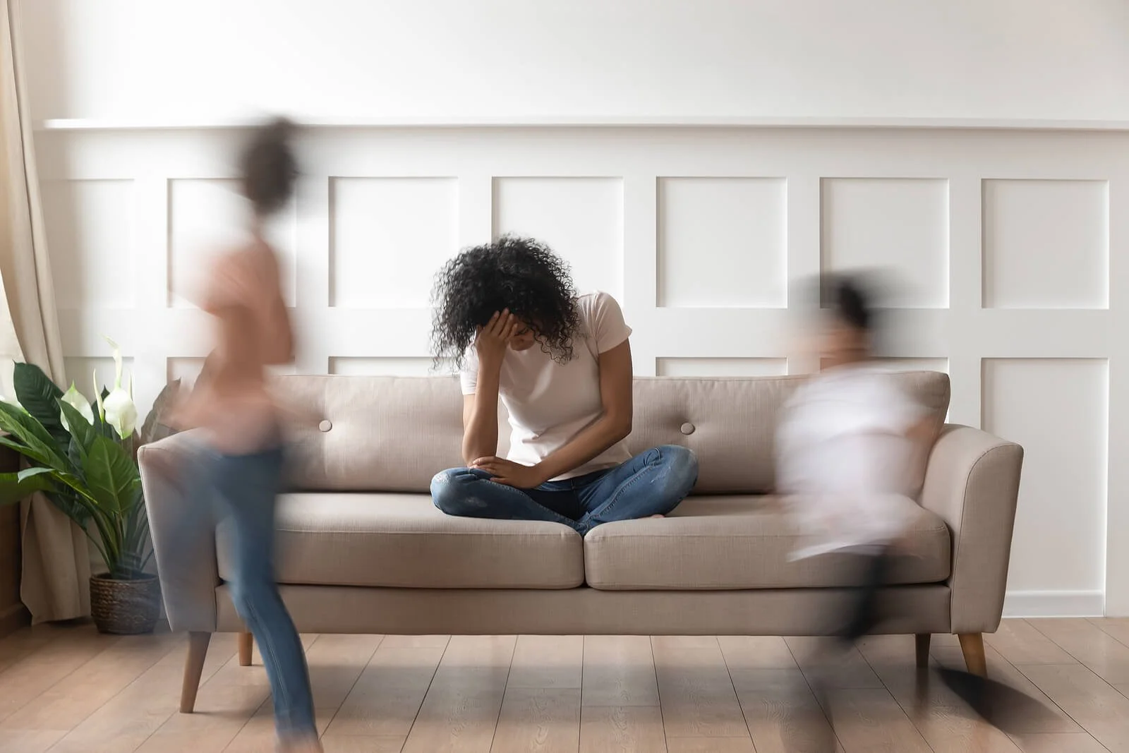 Parent sitting on a couch feeling overwhelmed while children move quickly around the room, representing listening and communication challenges at home