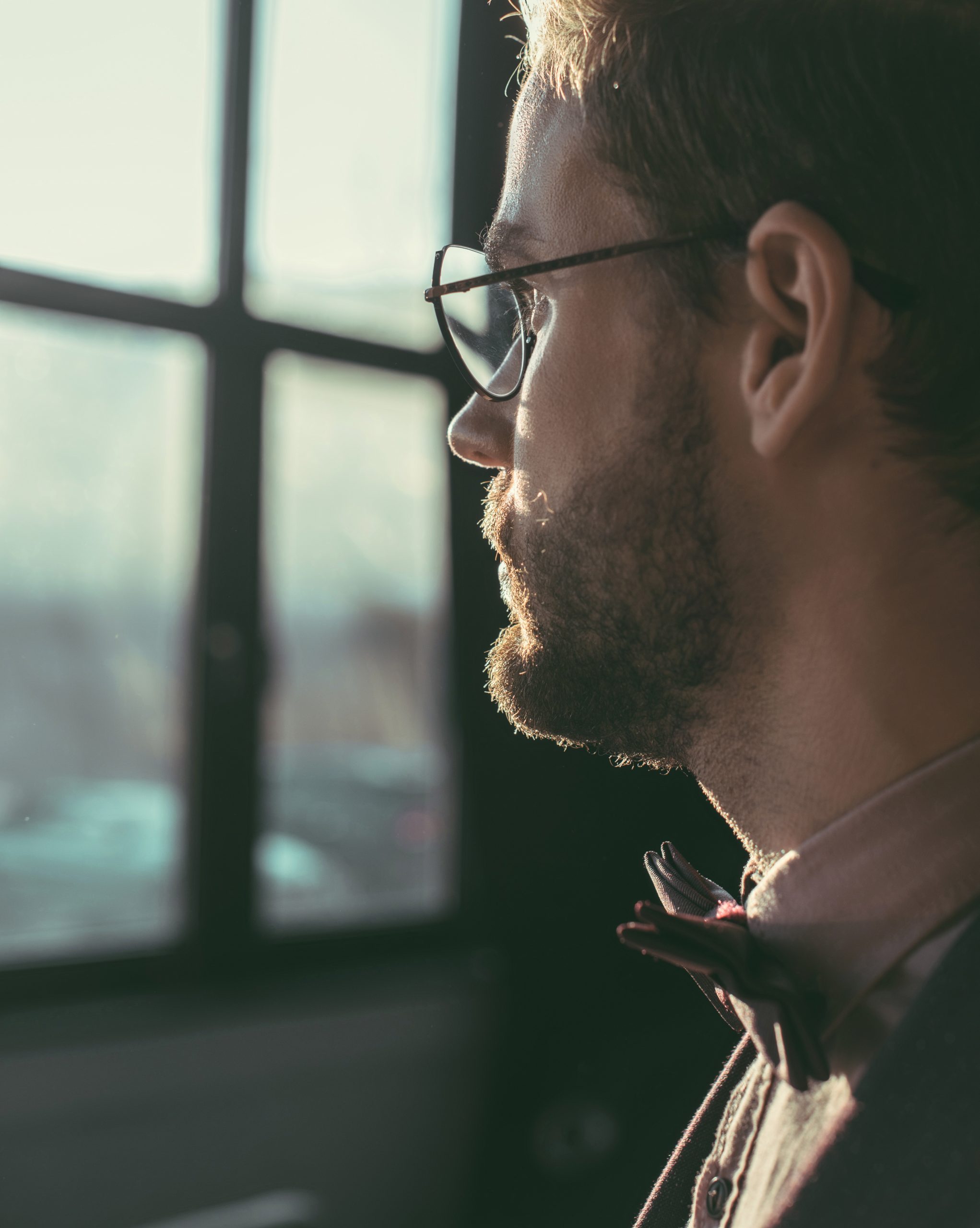 Man with glasses looking out a window with soft light on his face.