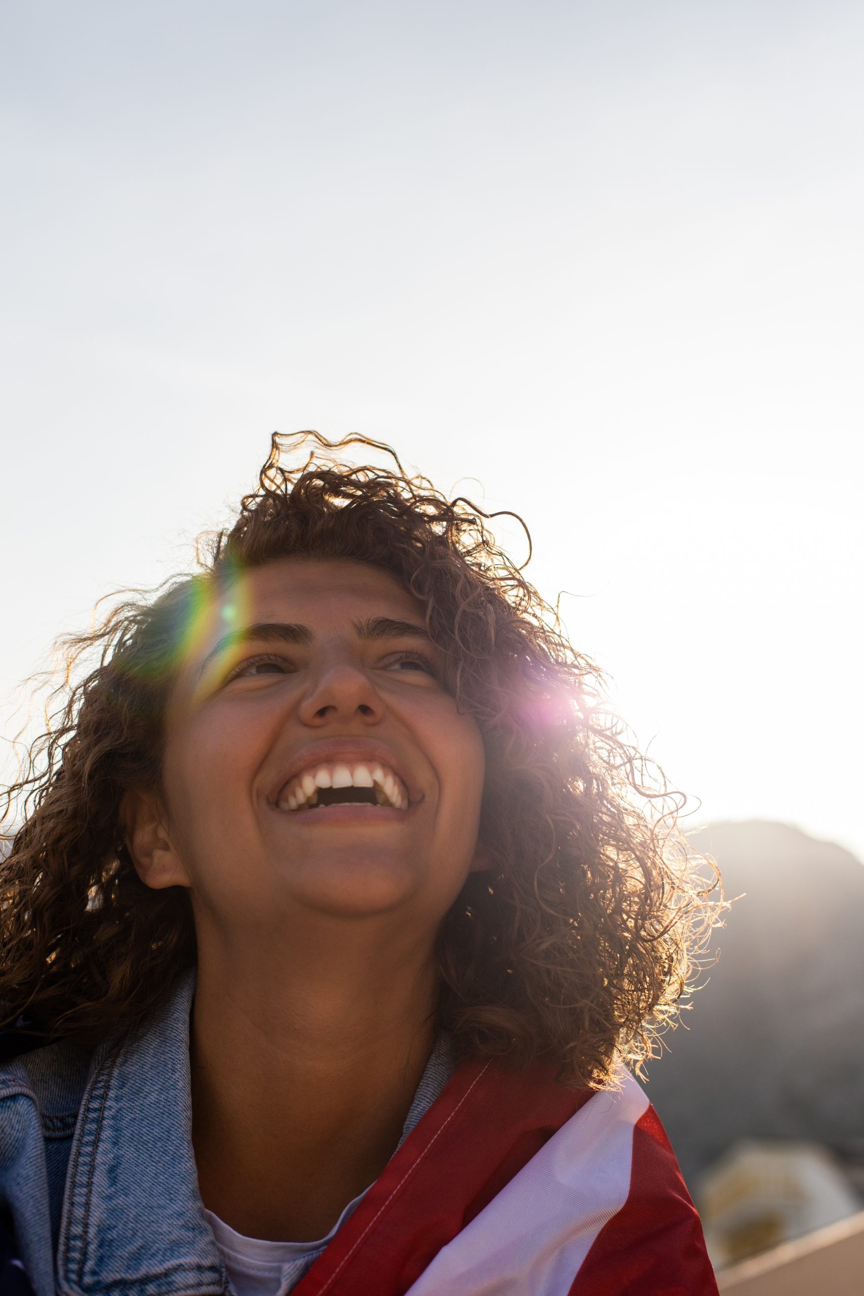 happy young woman with curly hair and flag wrapped around her, smiling up at the sun
