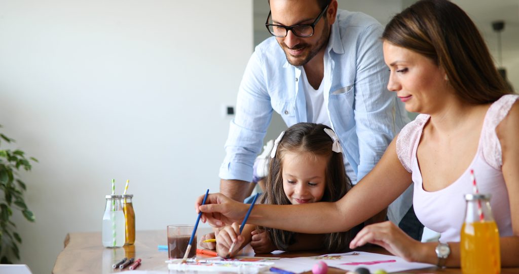 Family helping a young girl paint at a table together.