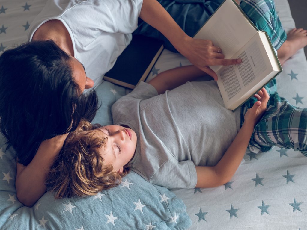Parent reading a book in bed beside a resting child.