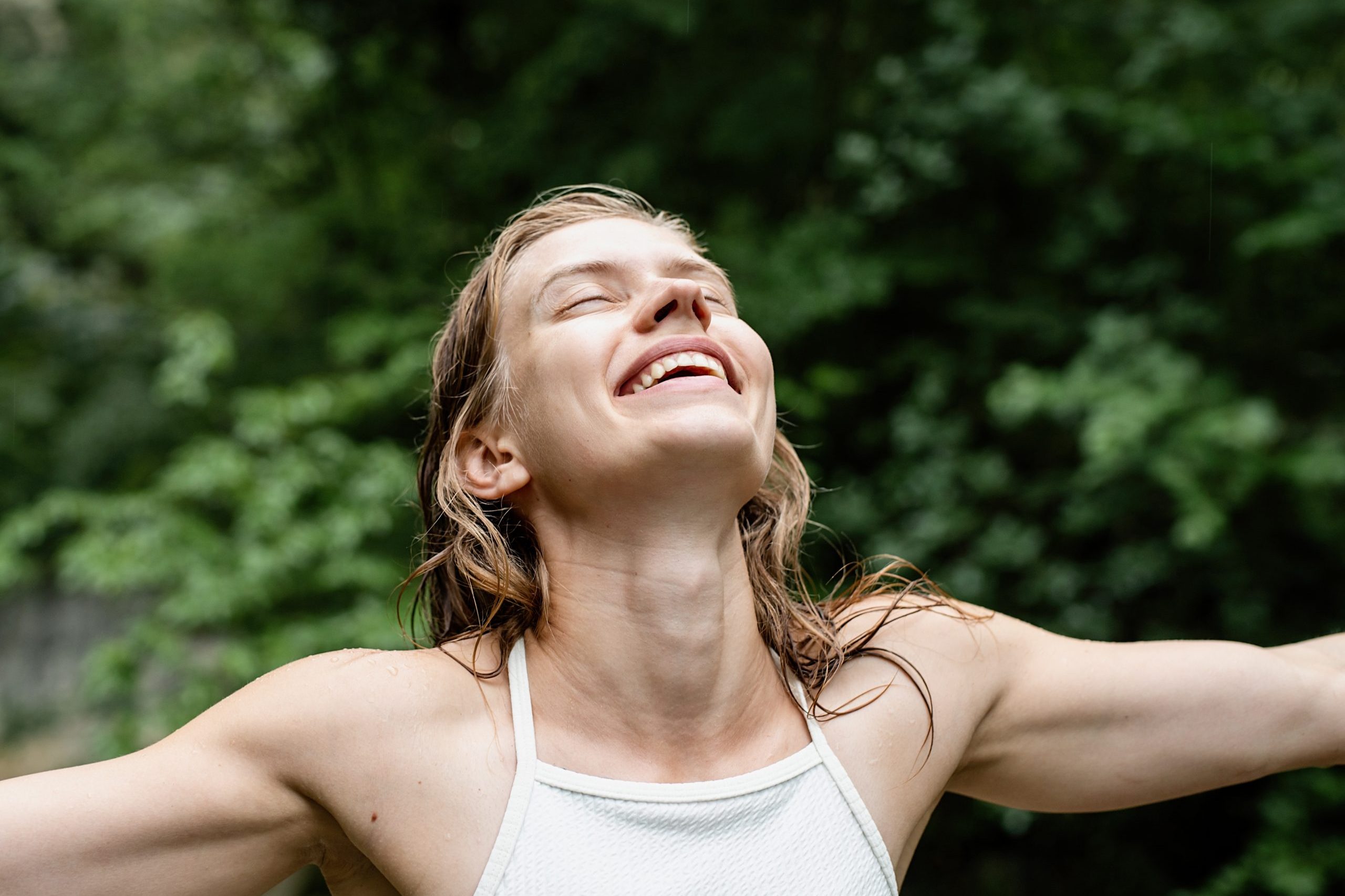 Woman turning her face to the sky and laughing, eyes closed. Green trees on the background
