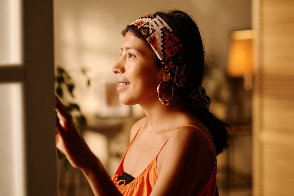 Side view of young smiling Latino woman in headscarf looking through window while standing in front of camera in living room