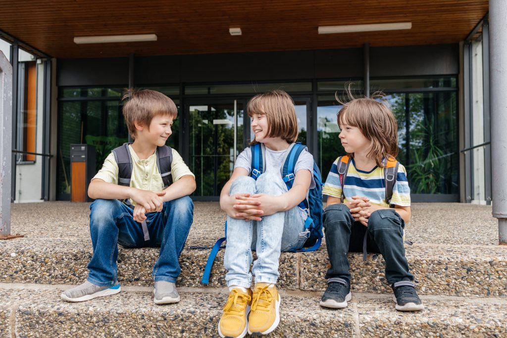 Three school-age children with backpacks sitting on steps and talking.