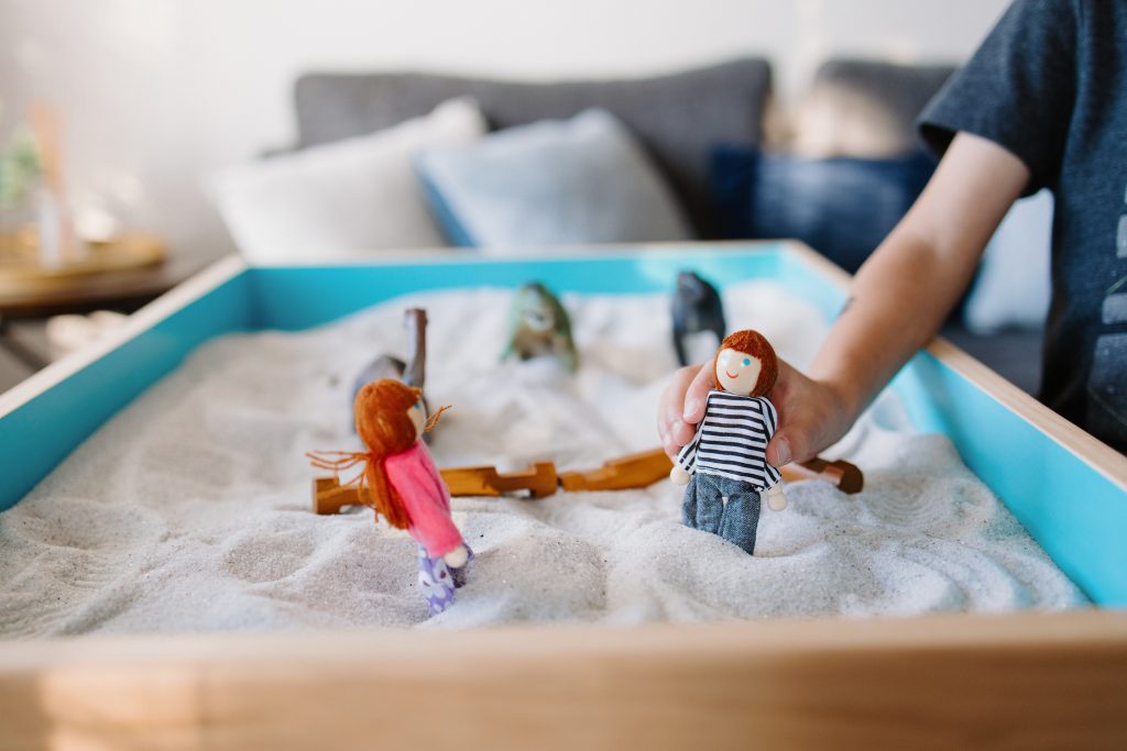 Child’s hand moving small dolls in a sand tray during play therapy.