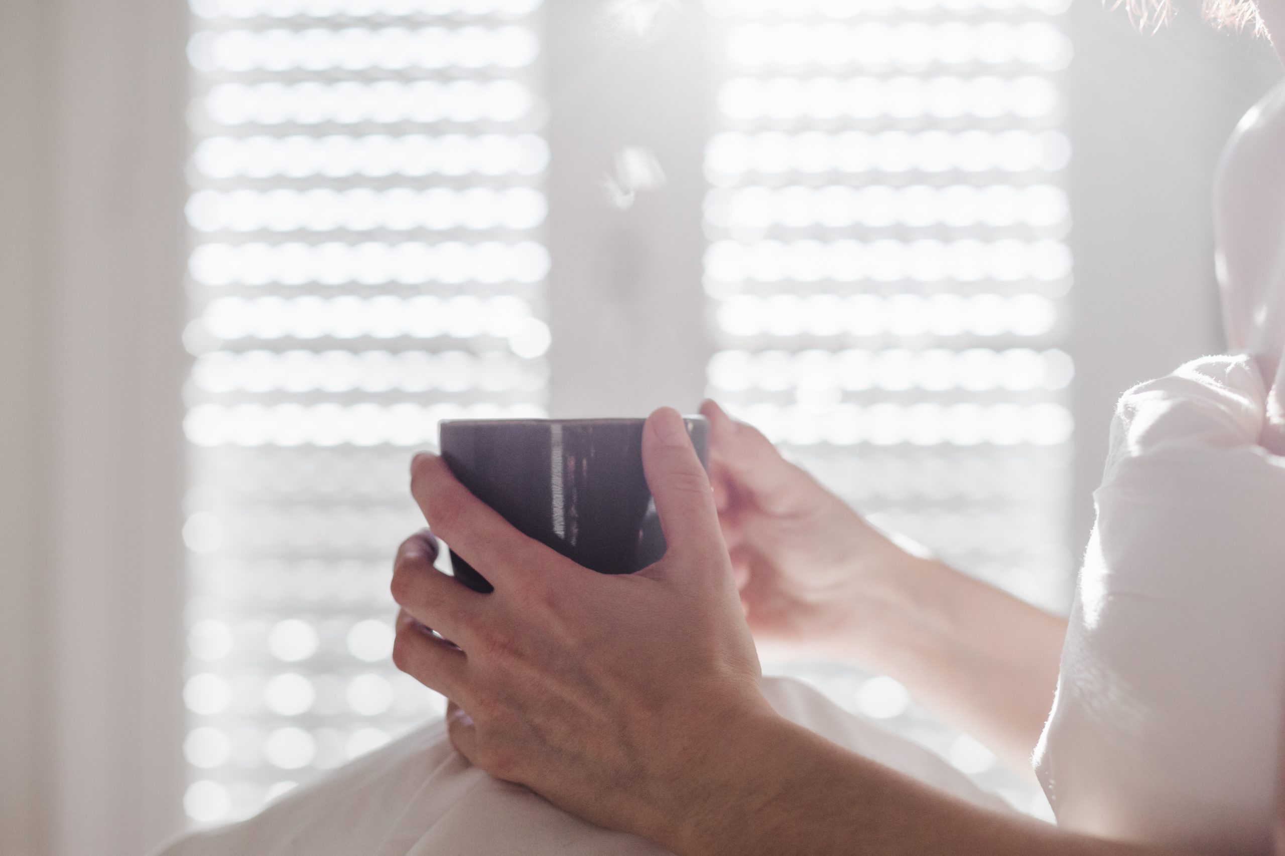 Hands holding a warm mug in front of a bright window.