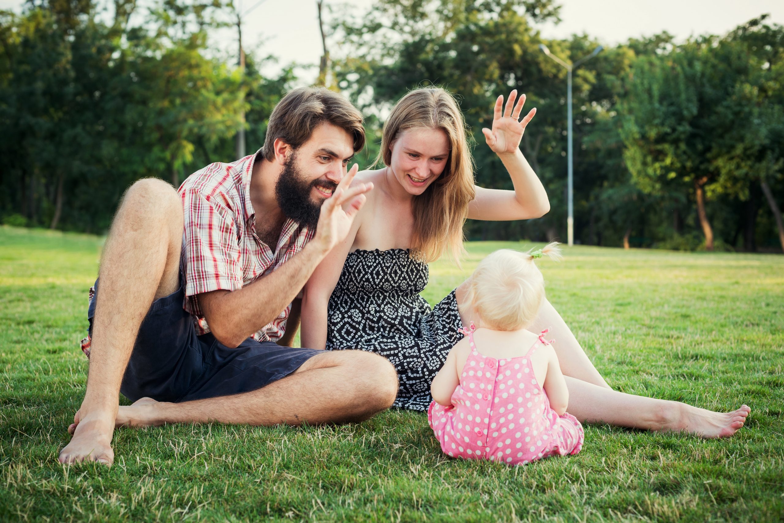 Parents sitting on the grass smiling and waving to their toddler.